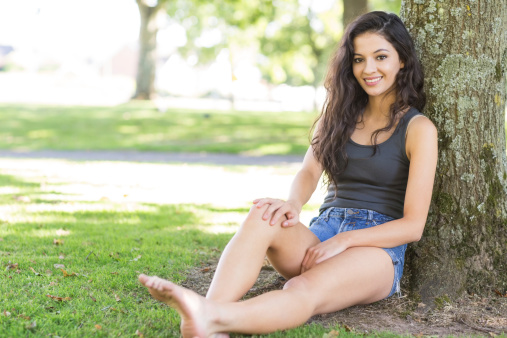 Casual attractive brunette sitting leaning against tree in a park on a sunny day