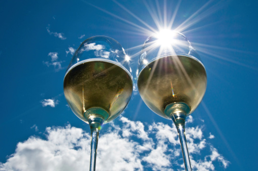 closeup of two wineglasses with a sun burst filled with white wine side by side with bright blue sky & clouds in the background