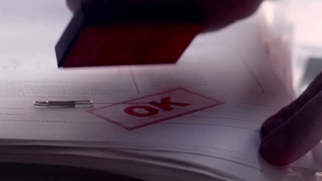 Close up, slow motion dolly video clip of a man’s hand rubber stamping a paper document with a red inked “OK” stamp. The documents have passed the grade; maybe a successful application; efficient work practise or simply completed clerical work.