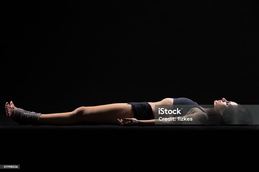 Savasana Yoga Pose Beautiful cool young fit woman in sportswear lying in Shavasana (Savasana, Corpse or Dead Body Posture), resting after practice, meditating, breathing, side view, studio, black background Shavasana Stock Photo