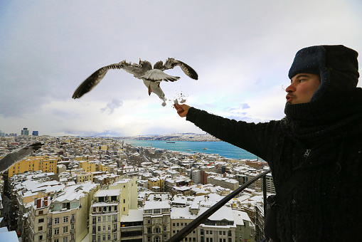 Kalpak wearing man feeding seagulls in Galata Tower on snowy landscape