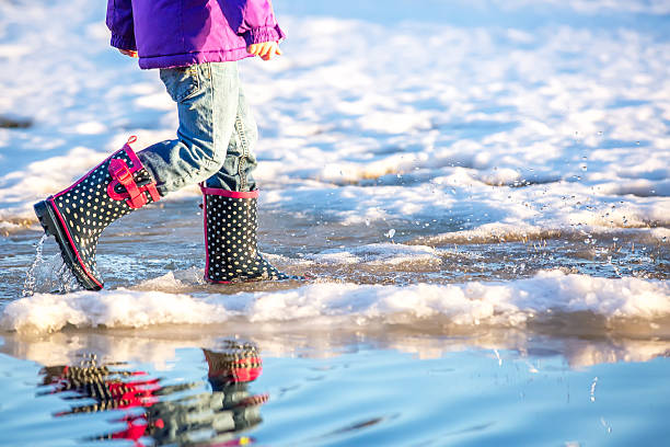 Waist down side view of a young girl walking through a large puddle of melted snow on an early springtime evening. The bottom of a purple jacket is visible, as well as light denim blue jeans, and navy blue with white polka dots and pink trim rubber boots.