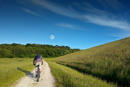 A young man is riding a bike in the early evening, at Bewl Water in East Sussex. There is a full moon. The reservoir dam can be seen, rising up to the right hand side of the cyclist.