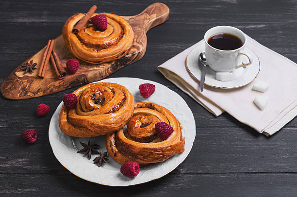 Round twisted Denish Buns Round twisted Denish Buns with berries raspberries, syrup, white cup of espresso coffee, sugar cubes, cinnamon sticks, star anise, cutting board made of wood olive on a black wooden background denish stock pictures, royalty-free photos & images