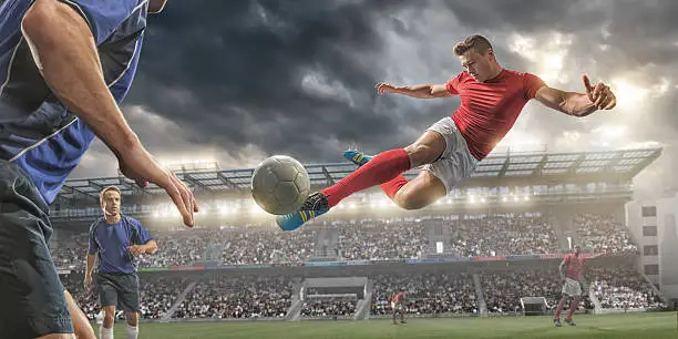 A close up image of professional soccer player jumping in mid air, about to volley football past rival players during a soccer game. Action takes place in a generic outdoor floodlit stadium full of spectators under a dramatic stormy sky at sunset. All players are wearing generic unbranded football kit. 