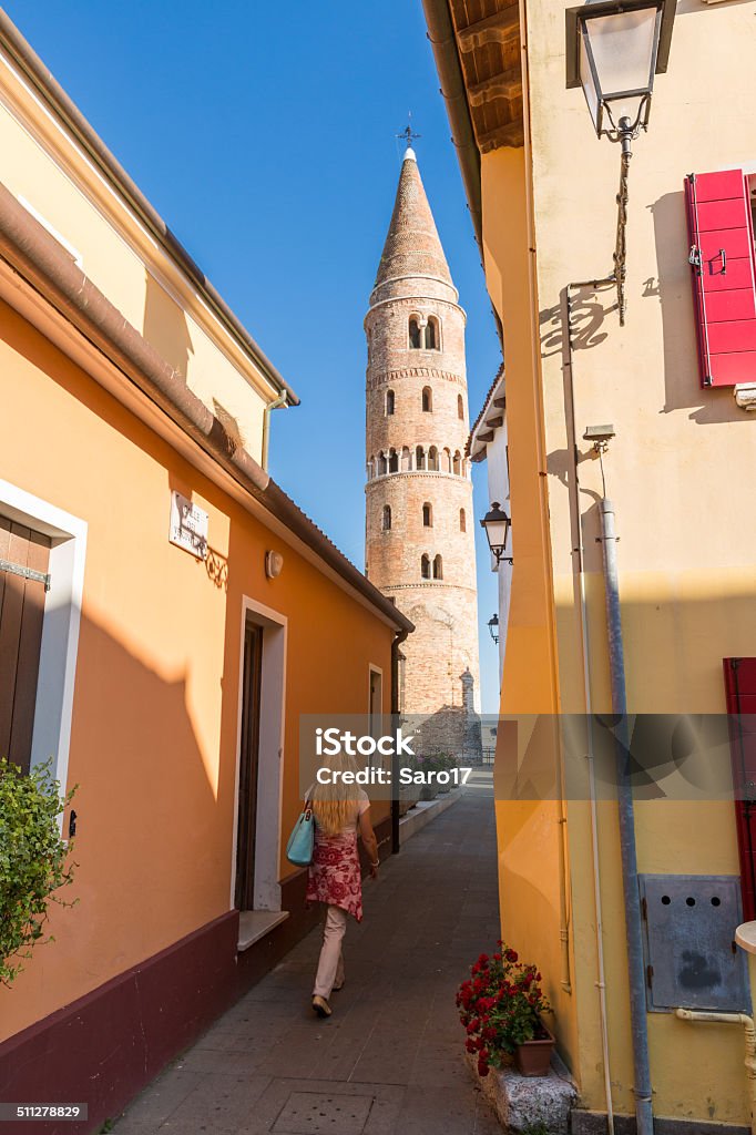 Exploring Caorle Old Town, Italy A blonde haired woman is exploring the Old Town of Caorle. Caorle is a coastal town in the province of Venice, Veneto, Italy, located between the estuaries of the Livenza and Lemene rivers. It is situated on the Adriatic Sea. Caorle is a small city, but is frequented by tourists in all seasons. The old town centre is still structured as a little Venetian corner, with its coloured houses, its "calli" (alleys) and "campielli" (squares). In the Background  the characteristic bell tower, dating to 1048, rises to a height of 48 meters. It is a typical example of Romanesque style, but it has a cylindrical structure, and it is surmounted by a cone-shaped cusp, that makes it unique in the world. Adult Stock Photo