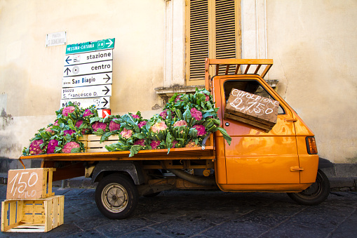 A cute orange vintage truck selling purple cauliflower on a street in Sicily, Italy. Yellow wall and shutters and road signs in the background.