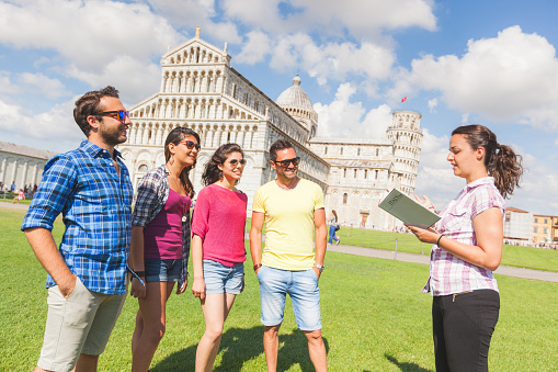 Group of tourists in Pisa, Italy. A group of friends is listening to a guide talking about a famous monument. They are two women and two men wearing summer clothes. They are a multicultural group on holidays.