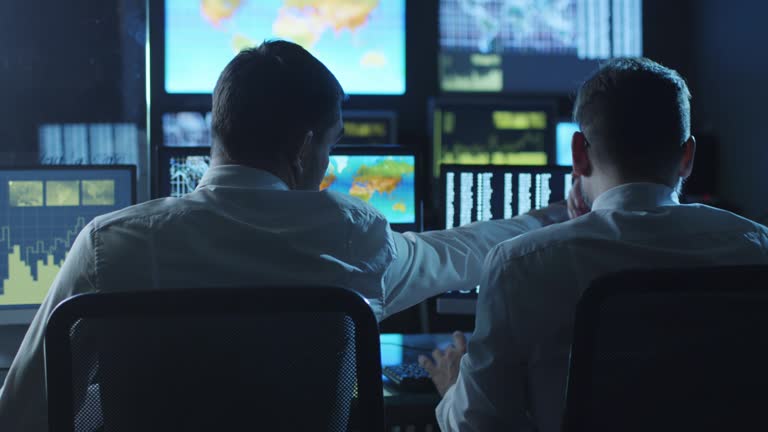 Two employees are working on computers in a weather forecast center filled with display screens.