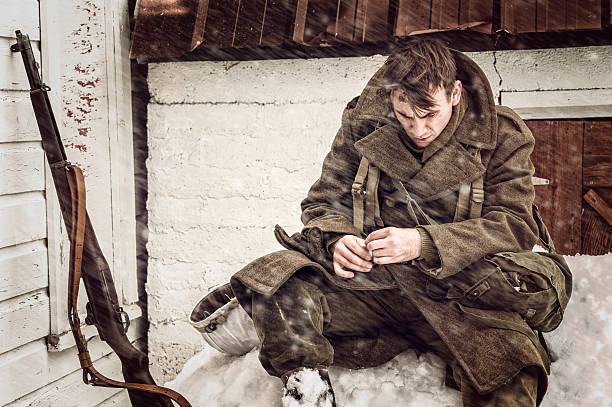 Tired and Cold WWII US Soldier Outdoors In a Blizzard.  He is looking down and has his rifle leaning against an old barn wall.  A blizzard is swirling all around him.  Longing for home