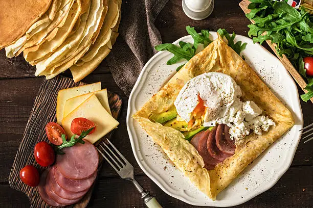 Crepe galette with meat, avocado, soft white cheese and poached egg on white plate. Sliced yellow cheese, pastrami, cherry tomatoes, green salad and stack of crepes on side. Top view. Food gasing