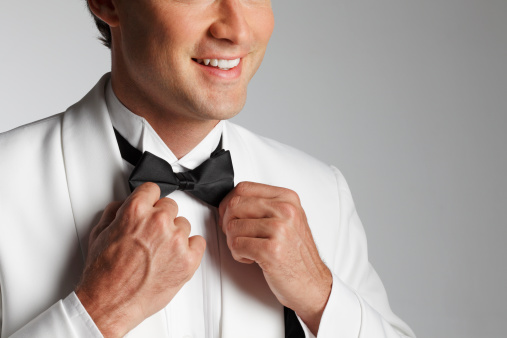 A close up of a man in a  white tuxedo adjusting his bow tie.