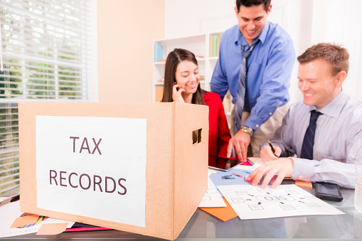 ]Cheerful, multi-ethnic firm of tax accountants work together on customer income tax returns.  Two business men and one woman.  Digital tablet, smart phones along with documents lie on desk.  Large box of tax records in foreground.