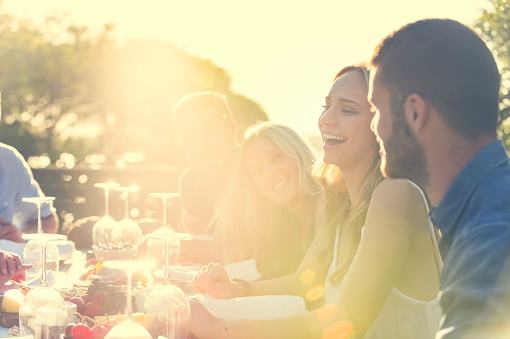 Group of young people eating outdoors. They are all happy, having fun, smiling laughing and talking. There are fruit and chees platters on the table and some salad. Back lit with lens flare.