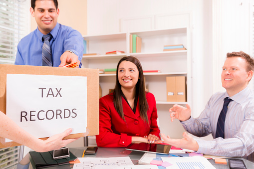 Cheerful, multi-ethnic firm of tax accountants work together on customer income tax returns.  A client brings in a box of tax records in foreground.  Two business men and one woman.  Digital tablet, smart phones along with documents lie on desk.