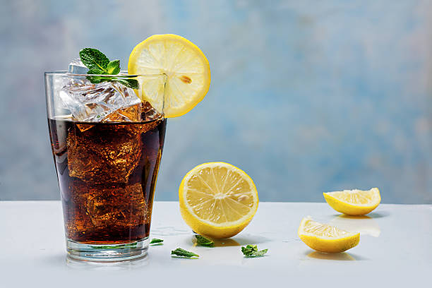 glass of cola  with ice cubes and lemon slices glass of cola or ice tea with ice cubes, lemon slice and peppermint garnish, against a blue wall non alcoholic beverage stock pictures, royalty-free photos & images