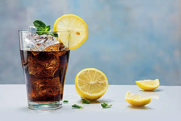 glass of cola or ice tea with ice cubes, lemon slice and peppermint garnish, against a blue wall