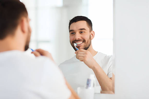 man with toothbrush cleaning teeth at bathroom health care, dental hygiene, people and beauty concept - smiling young man with toothbrush cleaning teeth and looking to mirror at home bathroom human teeth stock pictures, royalty-free photos & images