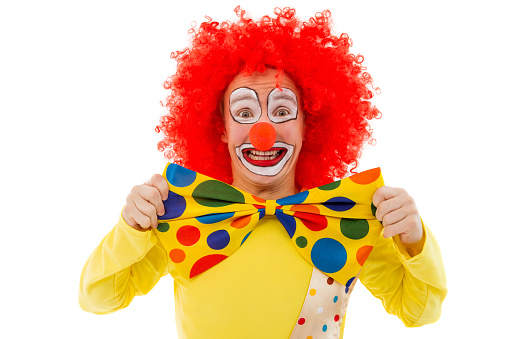 Portrait of a funny playful clown in red wig adjusting his big colorful bow tie, looking at camera and smiling, isolated on a white background
