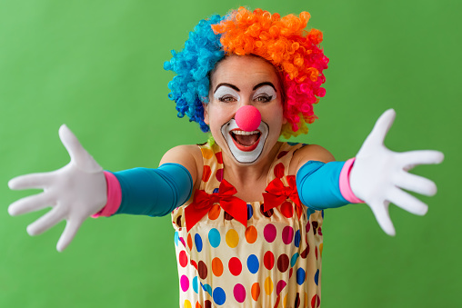 Portrait of a funny playful female clown in colorful wig stretching her hands like ready to hug and smiling, standing on a green background
