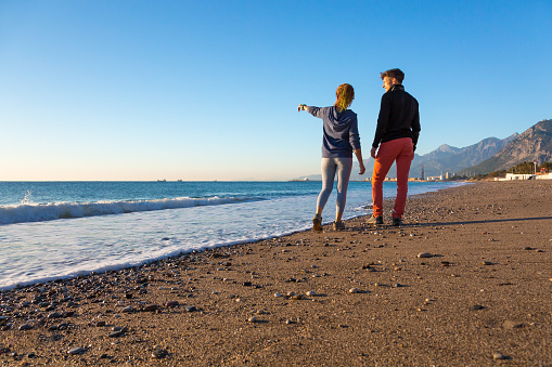 Man and Woman walking on Beach and enjoying Sea View Girl Pointing with hand on Vessels on Horizon and Sea surf