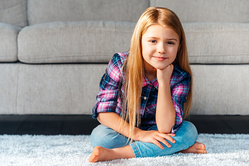 Cheerful little girl looking at camera with smile while sitting on the carpet in lotus position at home