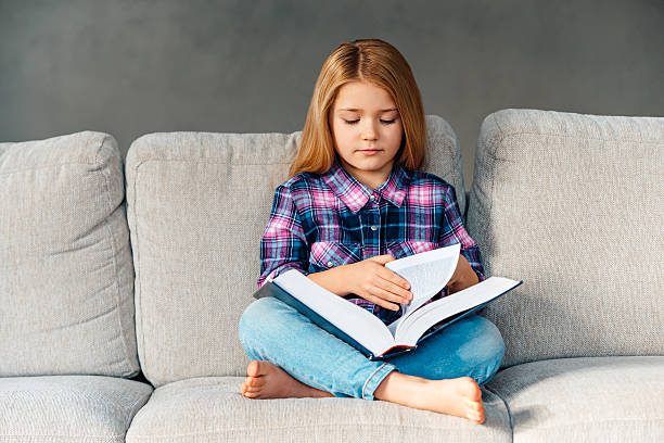 Time for fairytale. Beautiful little girl reading book while sitting on the couch in lotus position at home kid sitting cross legged stock pictures, royalty-free photos & images