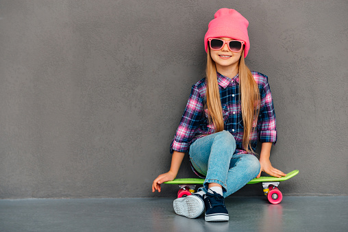 Full length of cheerful little girl in sunglasses looking at camera with smile while sitting on skateboard against grey background