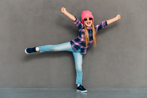 Full length of cheerful little girl in sunglasses keeping arms outstretched and looking at camera with smile while standing on one leg against grey background