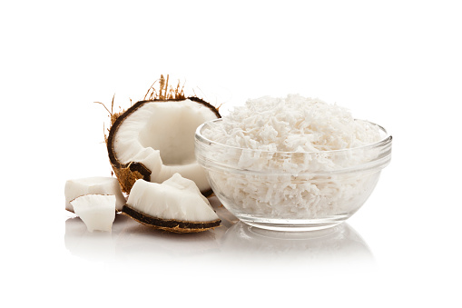 Glass bowl filled with grated coconut and half coconut with pieces isolated on reflective white background.