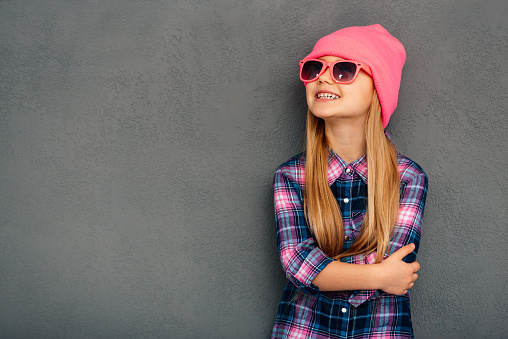 Cheerful little girl in sunglasses keeping arms crossed and looking up with smile while standing against grey background