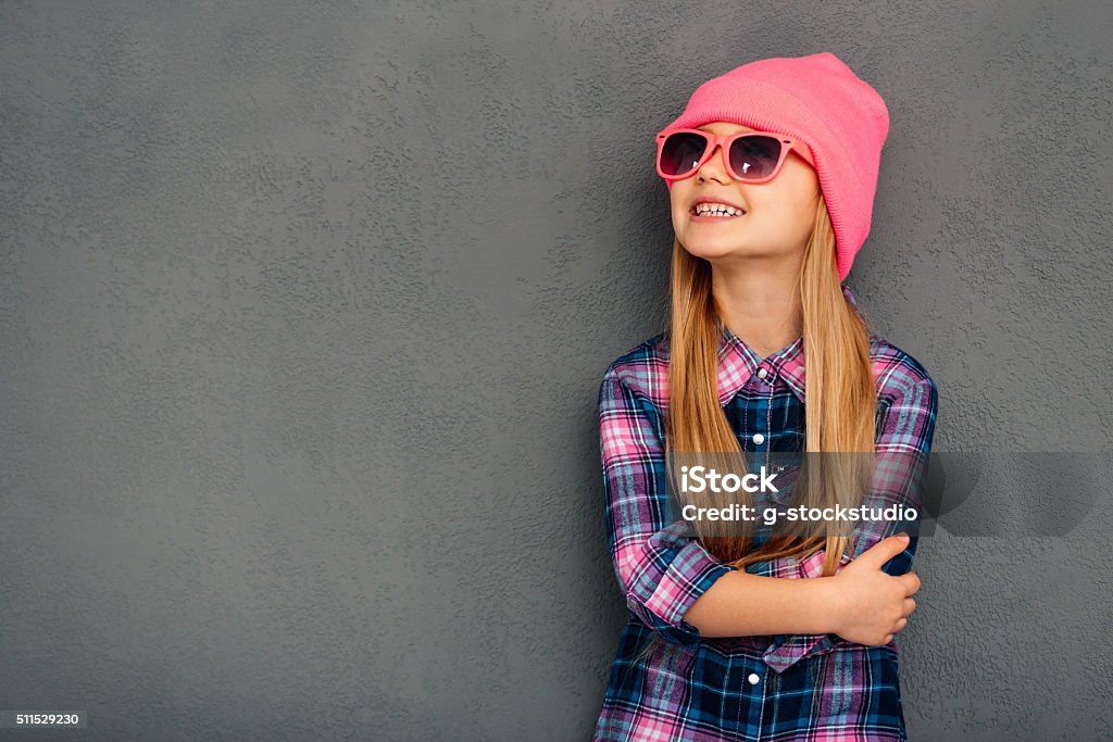 Stylish and cute. Cheerful little girl in sunglasses keeping arms crossed and looking up with smile while standing against grey background Child Stock Photo