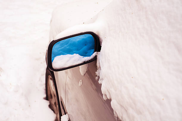 Close up of car covered by snow on snowy winter day. Reflection of fresh snow in automobiles mirror. Horizontal color image.