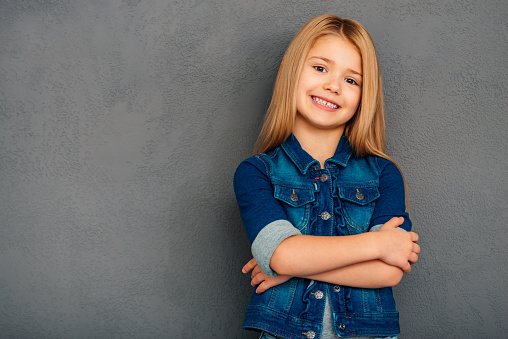 Cheerful little girl holding arms crossed and looking at camera with smile while standing against grey background