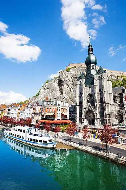 View of Collegiale Notre Dame de Dinant near Meuse river with white ship on it in Dinant, Belgium