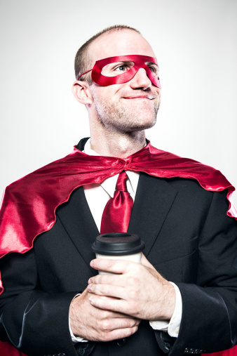 A business man in suit and tie wears a superhero cape and mask as well, a smile on his face as he holds his morning cup of coffee.  Vertical portrait.