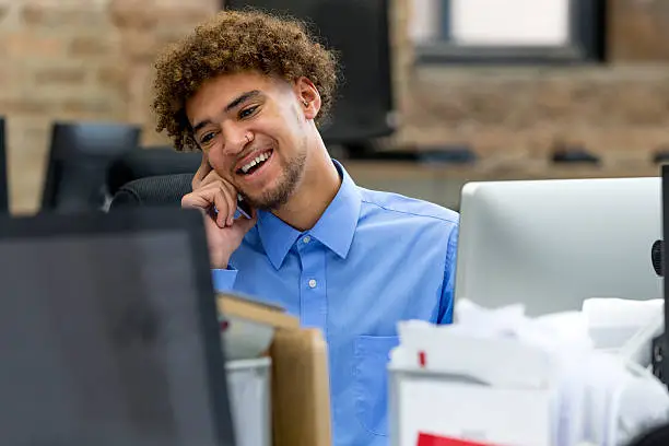 Business man on the phone smiling while sat in front of his computer screen. Close up of his head and shoulders off him sitting at his office desk. Positive emotions.