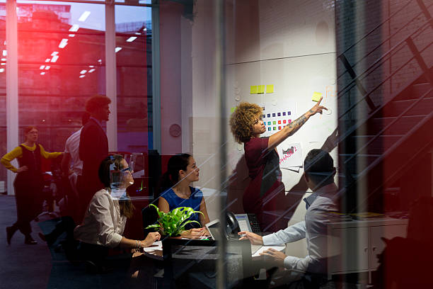 This is the business plan! Business woman dominates the meeting standing up giving instructions while other members sit looking and listening. Female woman is pointing to notes etc in front of her on the whiteboard. low lighting stock pictures, royalty-free photos & images