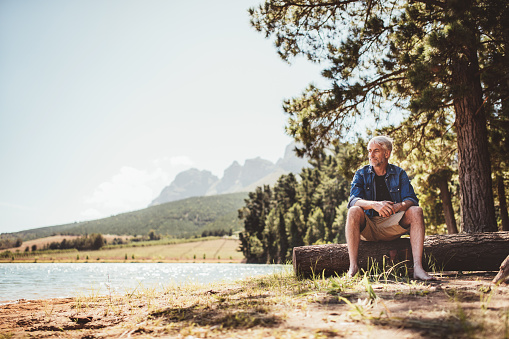 Horizontal shot of the mature man sitting on the wooden log by the lake. Senior man relaxing near a lake on a summer day.