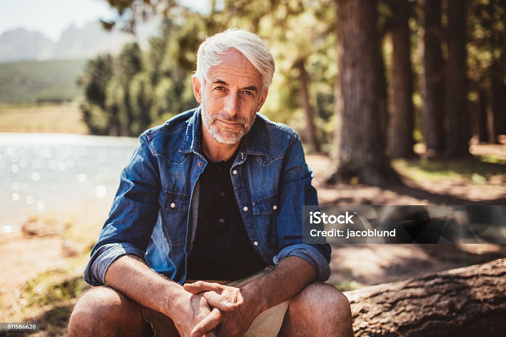 Senior caucasian man relaxing on a log by the lake Portrait of mature man sitting near a lake staring at camera. Senior caucasian man relaxing on a log by the lake on a summer day. Men Stock Photo