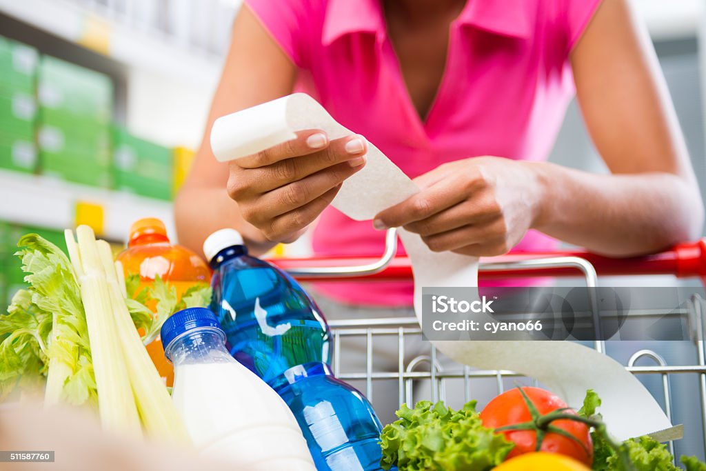 Expensive grocery bills Unrecognizable woman checking a long grocery receipt leaning to a full shopping cart at store. Budget Stock Photo