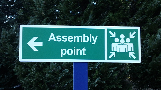 A green sign with white text saying 'Assembly Point' and an arrow pointing to the left. Also a white standard ISO pictogram indicating the same. A safety organisation sign.