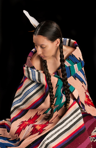 An American Indian young woman is sitting while wrapped in a blanket. She looks downward and wears a somber facial expression. Her long hair is braided with a feather in her hair.