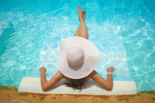 beautiful woman in a white hat sitting on the edge of the pool.