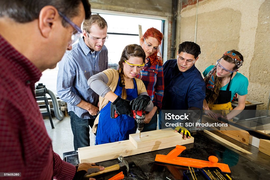 Woman drills hole in board in makerspace Young Hispanic woman uses a power drill to drill a hole into boards. Mature Caucasian manager and diverse team of male and female employees gather around her as she shows them how to use a drill. Carpenter Stock Photo