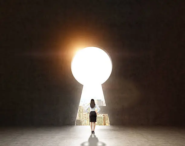 woman with hands on hips standing in front of a huge keyhole, city and sky seen through it. Black background. Back view.  Concept of finding the way