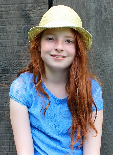 Photo showing a young girl sitting on a stone garden bench with long red hair, wearing a hat and a blue lace dress, and pictured smiling, looking forwards and showing how happy she is.  In the background is the side of a garden shed, made with wooden slats / planks of wood.