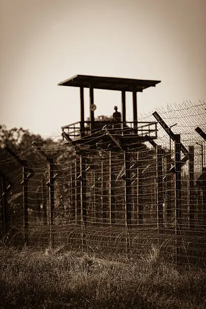 Watchtower, from "Coconut tree prison"  - a prisoner of war camp,  from Vietnam war in Phu Quoc.  Sepia, bokeh, vignette, vertical, perspective as seen from the main road. Focus on the fence's barbwire.