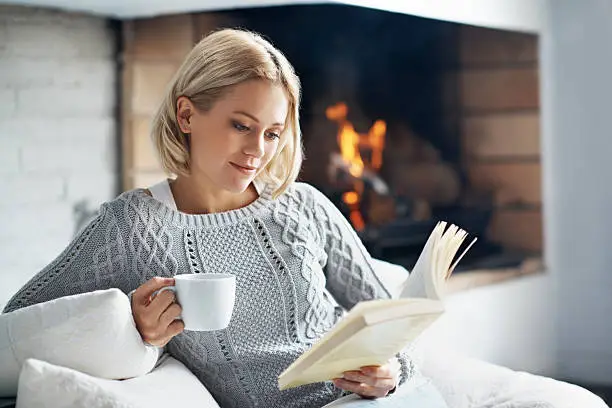 A beautiful young woman reading a book and enjoying a warm beverage near a fireplace