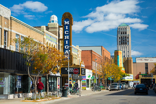 Ann Arbor, United States - October 18, 2015: People walking in the sidewalk of Liberty Street in Downtown Ann Arbor, with storefronts and the Michigan Theater sign, State Theater sign, cars and parked bicycles in the scene, and the Burton Memorial Tower in the distance, during a day with a blue sky with clouds.